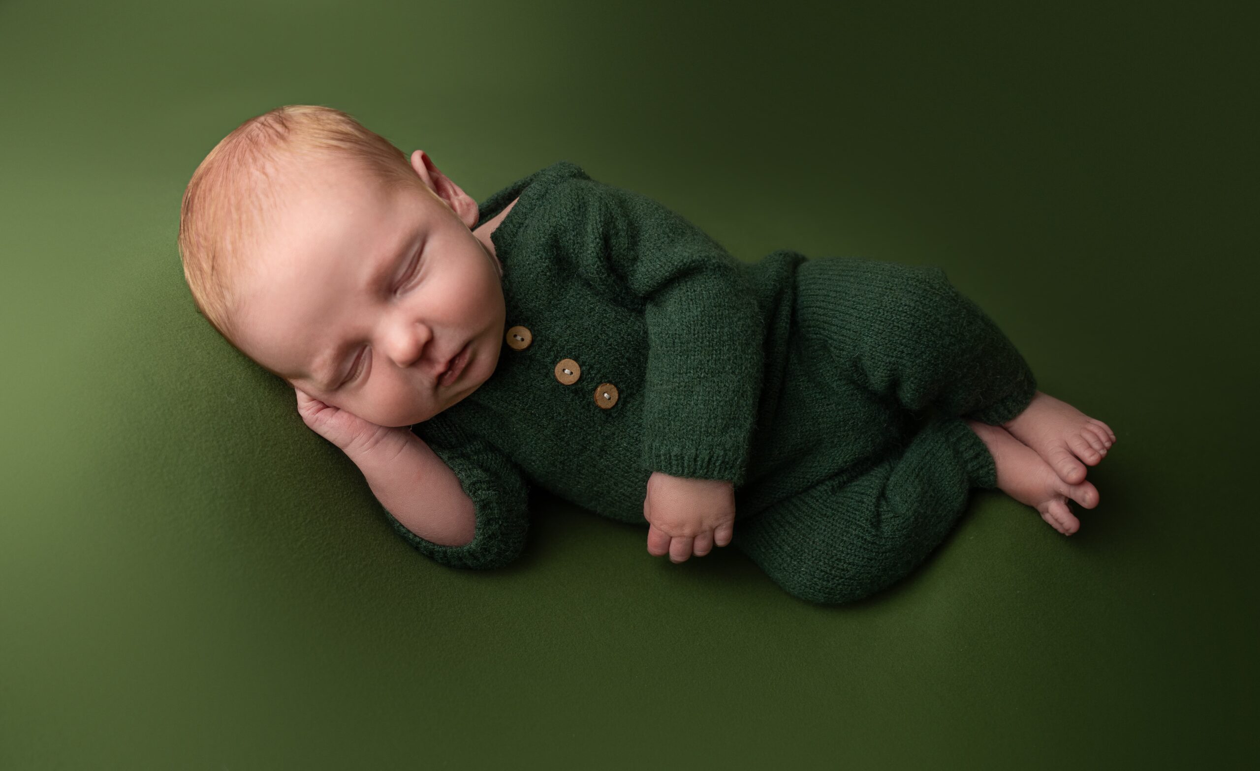 newborn baby boy posed in side laying pose on green backdrop in knit green onesie during in home newborn session with raleigh newborn photographer